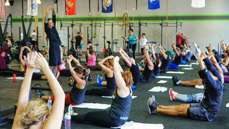 Hier siehst du einige Sportler/-innen bei einer Übung. Sie sitzen mit gestreckten Beinen auf dem Boden und haben die Arme grade nach oben gestreckt.