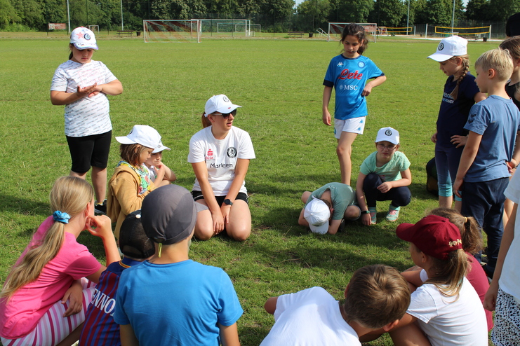 Hier siehst du eine Kindergruppe, die in einem Kreis auf einem Sportplatz versammelt sind.