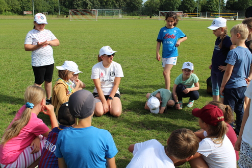 Hier siehst du eine Kindergruppe, die in einem Kreis auf einem Sportplatz versammelt sind.