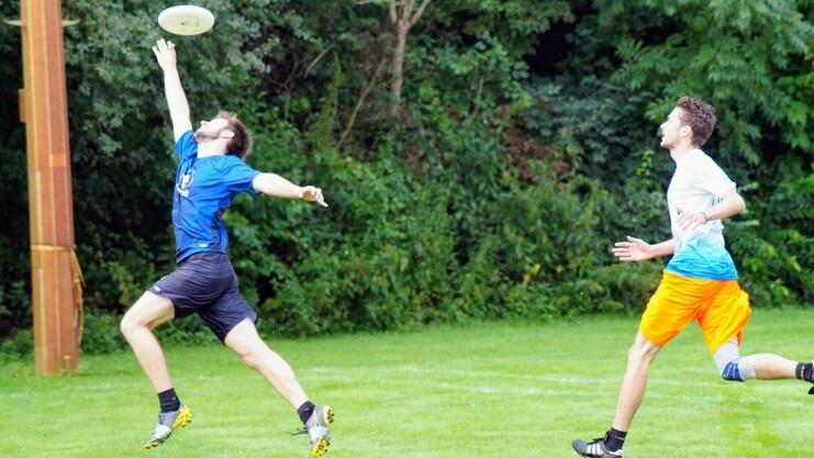 Hier siehst du zwei Männer. Der vordere springt und fängt eine Frisbee, der hintere läuft hinterher.