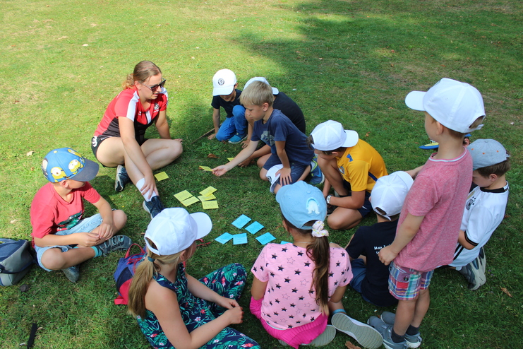 Hier siehst du eine Gruppe an Kindern im Kreis auf einer Wiese sitzen und ein Kartenspiel spielen. Sie tragen alle kurze Sachen und ein Cap.