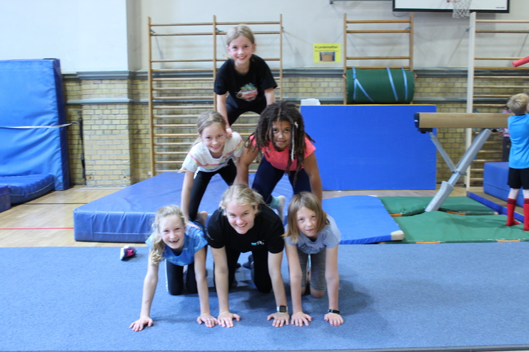 Hier siehst du sechs Kinder in einer Turnhalle. Sie machen eine Pyramide auf allen Vieren, bei edr unten drei Kinder sind, darüber zwei und oben eins.