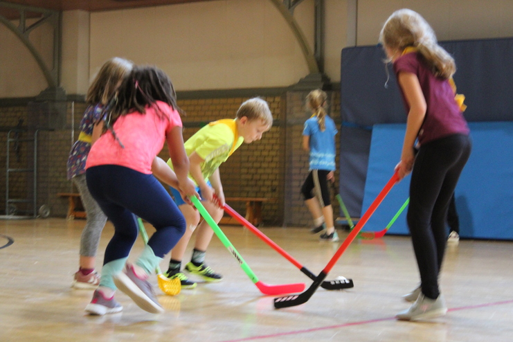 Hier siehst du Kinder in einer Turnhalle beim Unihockey spielen.