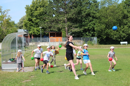 Hier siehst du eine Gruppe an Kindern draußen auf einer Wiese im Sommer. Sie haben alle einen Ball in der Hand und laufen.