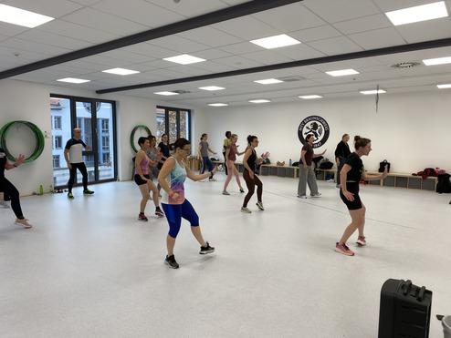 Hier siehst du den Kurs Jamfya. Dies ist eine Mischung aus Fitness und Tanzen. Auf dem Bild ist ein Kurs zu sehen. Sie haben die Arme leicht erhoben und tanzen.