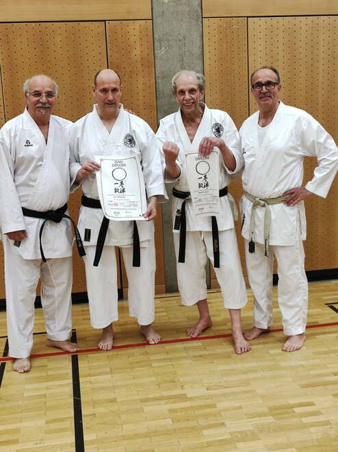 Hier siehst du vier Männer, in weißen Karateanzügen. Die Männer stehen nebeneinander und die beiden mittleren halten eine Urkunde in der Hand.