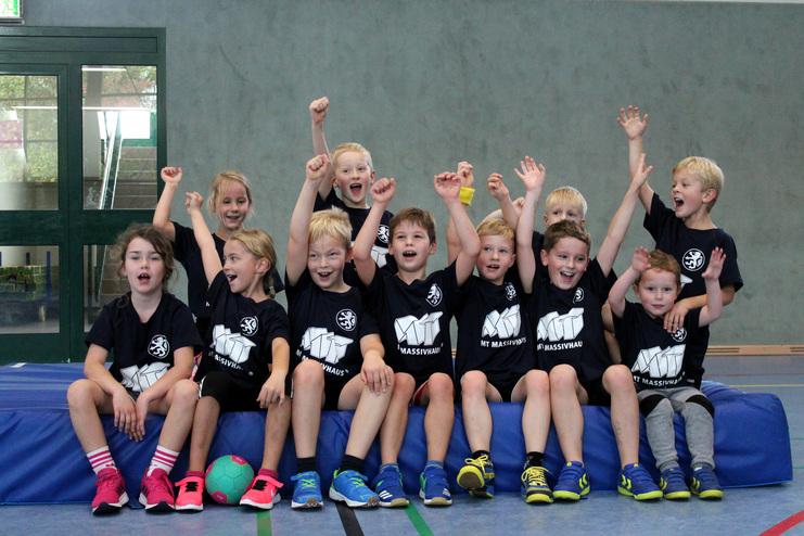 Hier siehst du ein Bild von vielen Kindern, die auf einer Matte sitzen und jubeln.