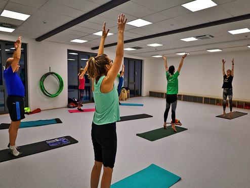 Hier siehst du einen Kurs. Mehrere Teilnehmend stehen auf der Matte und haben die Arme über den Kopf erhoben.