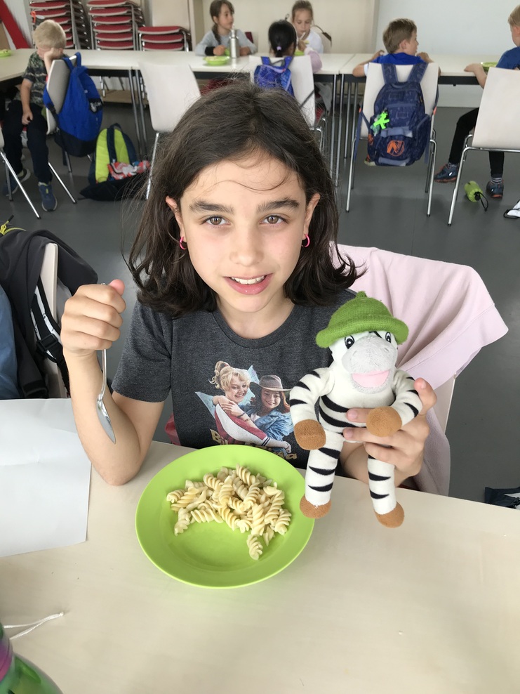 Hier siehst du ein M&auml;dchen auf einem Stuhl an einem Tisch sitzen. Es guckt in die KAmera, hat einen Teller mit Nudeln vor sich und in ihrer linken Hand eine Pl&uuml;schzebra.