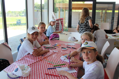 Hier siehst du eine Gruppe an Kindern am Tisch sitzen und malen. Sie gucken alle in die Kamera.