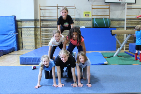 Hier siehst du sechs Kinder in einer Turnhalle. Sie machen eine Pyramide auf allen Vieren, bei edr unten drei Kinder sind, dar&uuml;ber zwei und oben eins.