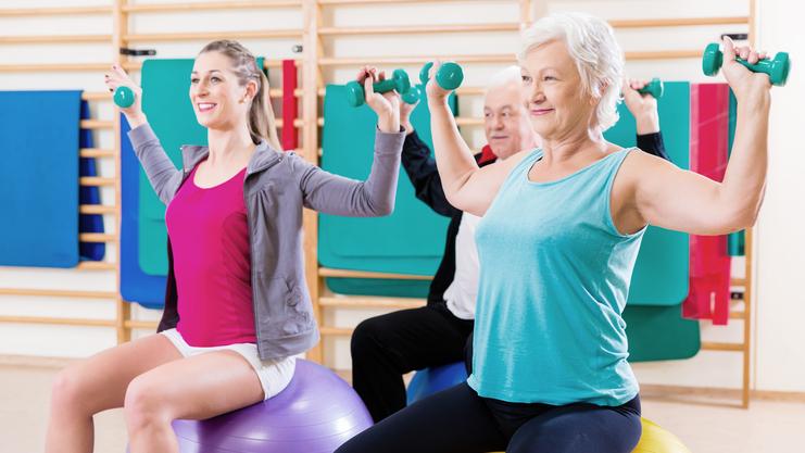 Hier siehst du drei Damen auf einem Pezziball sitzen und Gewichte in der Hand haben.