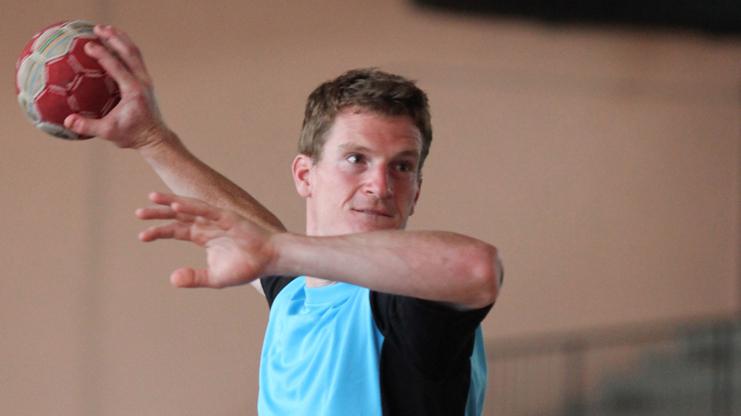 Handballspieler in blauer Sportkleidung wirft einen Ball in einer Sporthalle.