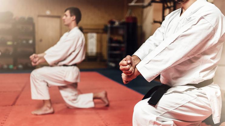Zwei Karateka in Trainingsanzügen knien auf einer Matte und üben eine Haltung. Der Raum ist mit Sportequipment ausgestattet.