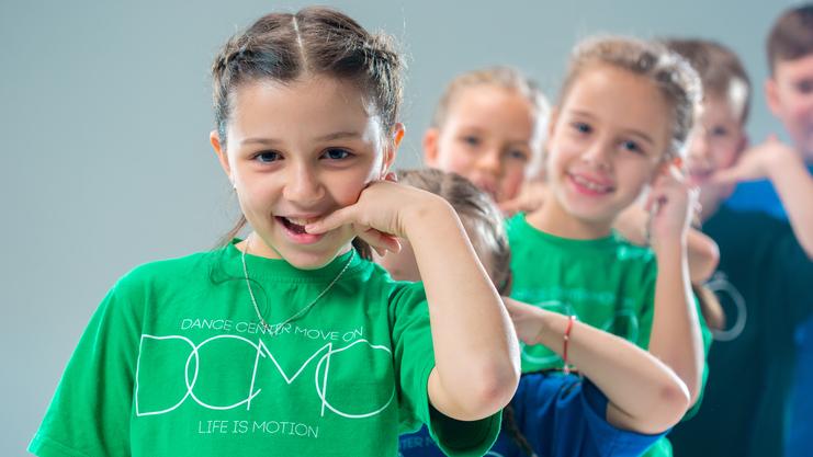 Lächelnde Kinder in bunten T-Shirts machen eine Tanzbewegung vor einem hellen Hintergrund.
