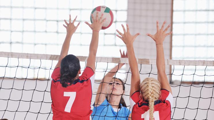 Zwei Spielerinnen in roten Trikots springen am Volleyballnetz, während eine dritte Spielerin auf den Ball zuspringt.